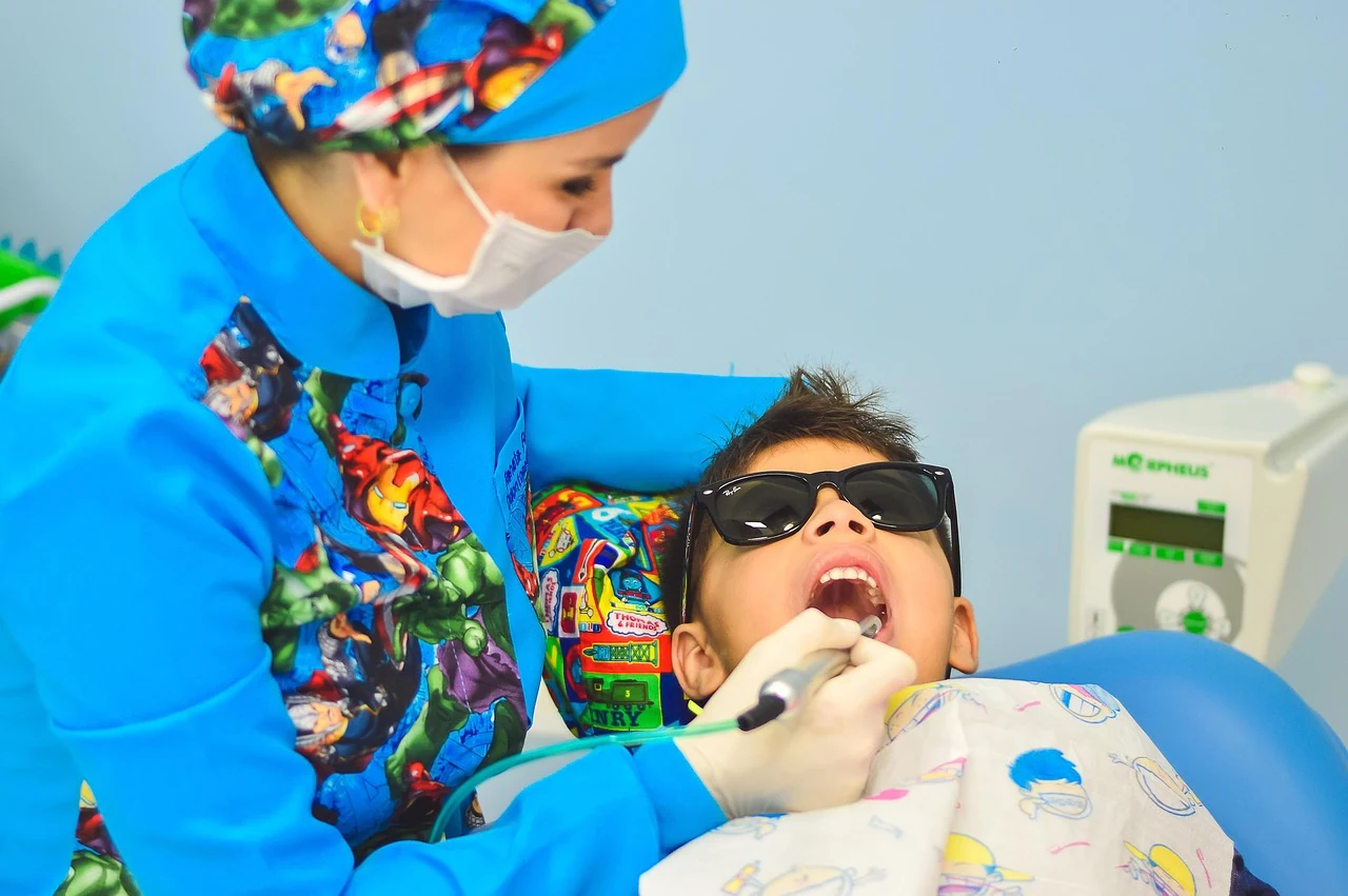 Dentist in a mask examines a boy's baby teeth in a dental chair, checking for cavities
