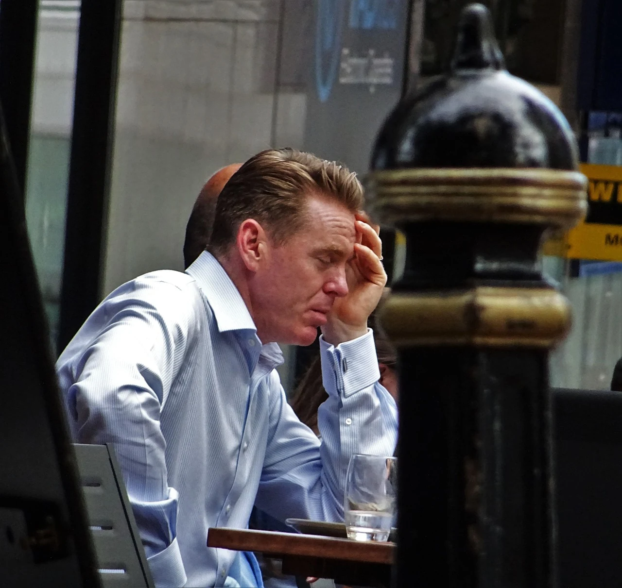 Tired man in a shirt sits at a café table with his head resting on his hand, showing chronic fatigue