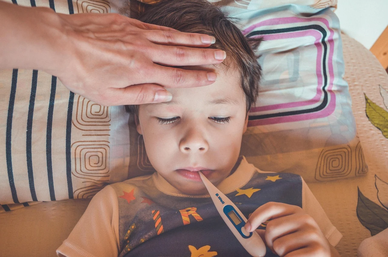 Adult hand resting on the forehead of a boy with a thermometer in his mouth, checking for fever in measles