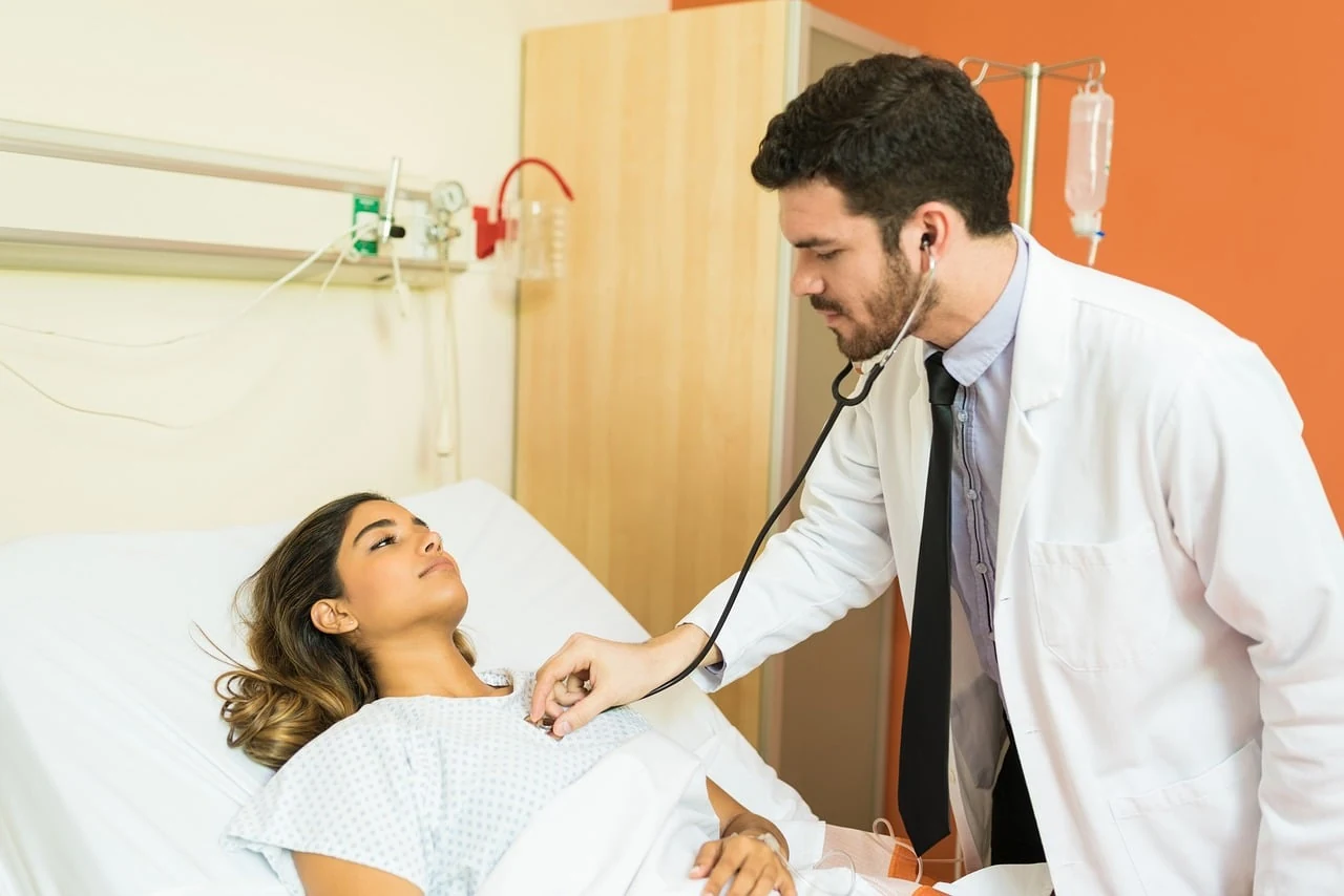 Doctor with a stethoscope examines a young woman lying in a hospital bed to find the cause of shortness of breath