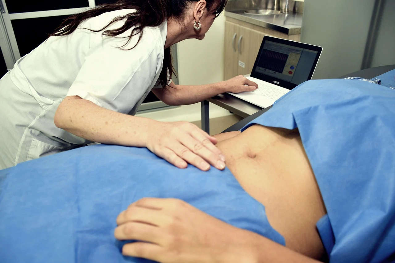 Doctor presses on a patient's abdomen under a blue drape while looking at a laptop, checking for gallstones