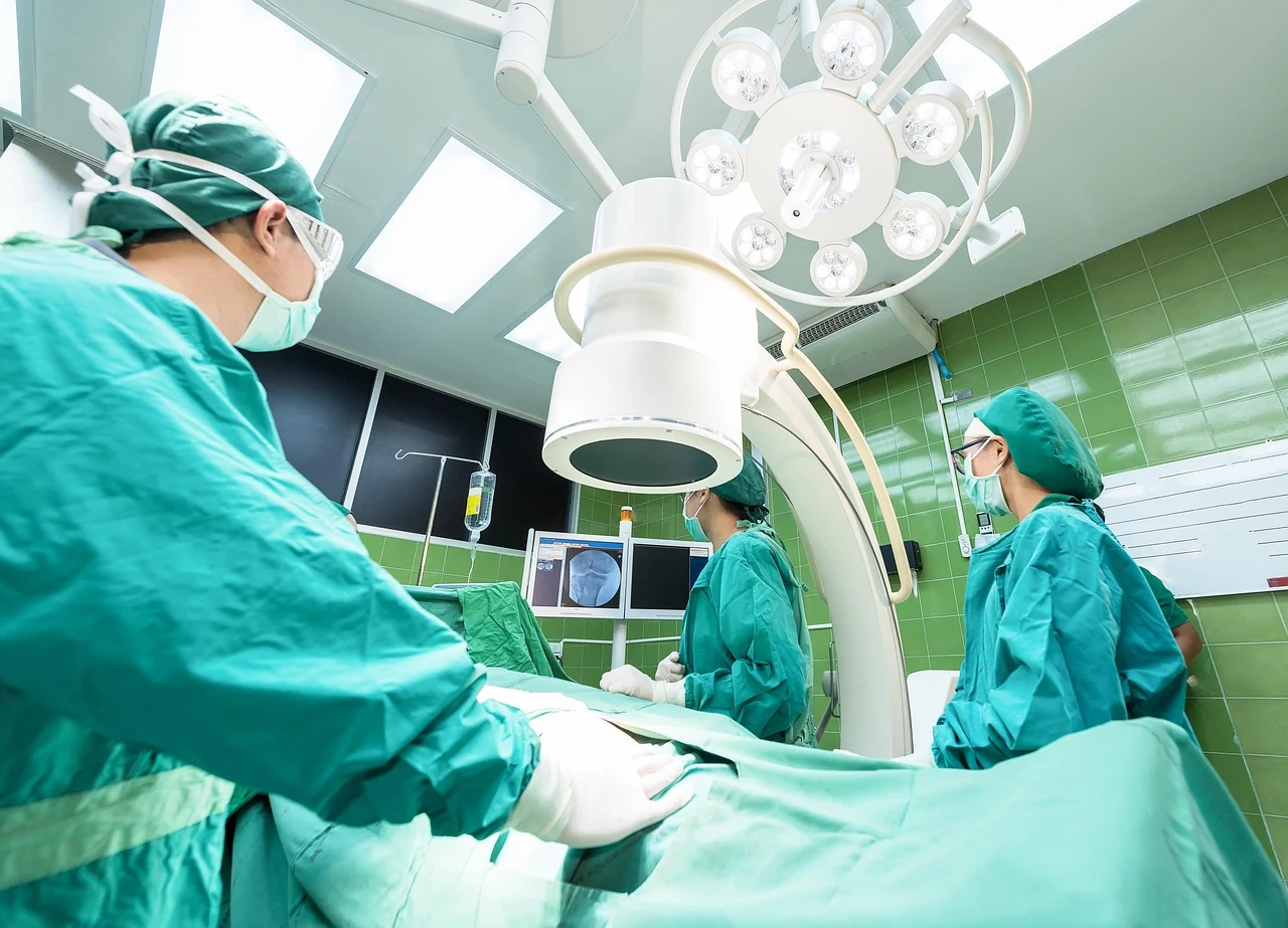 Surgeons in green scrubs and masks operate on a patient in an operating room under a large surgical light